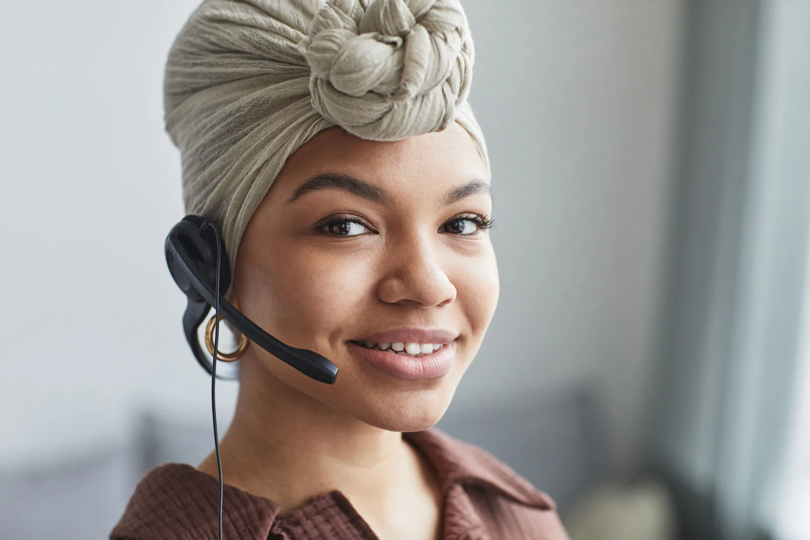 A smiling support agent in a cream headwrap wearing a headset, looking straight at the camera